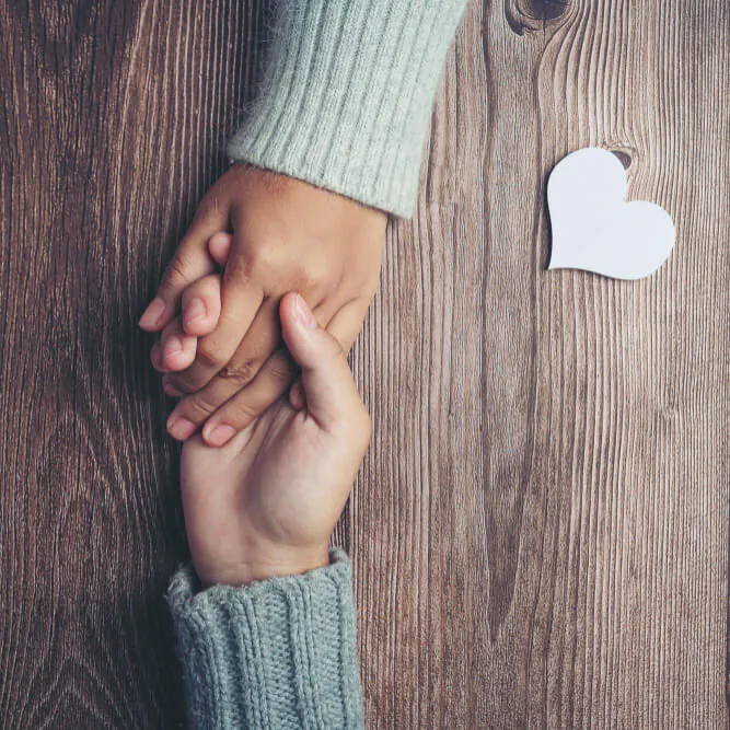 Two hands holding across a wooden table beside a paper heart