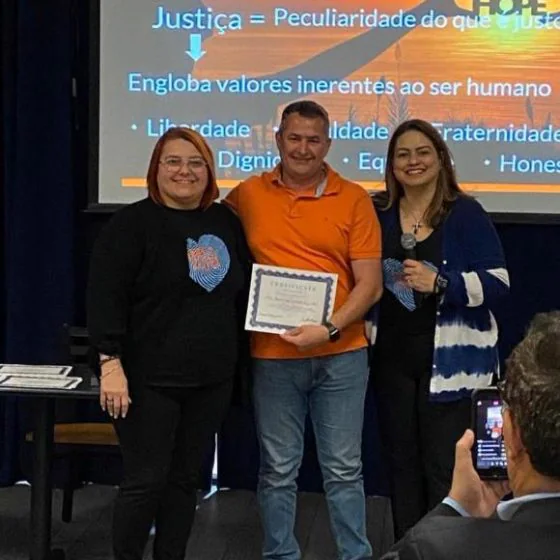 A participant receives his certificate between two organisers, slide on justice behind them